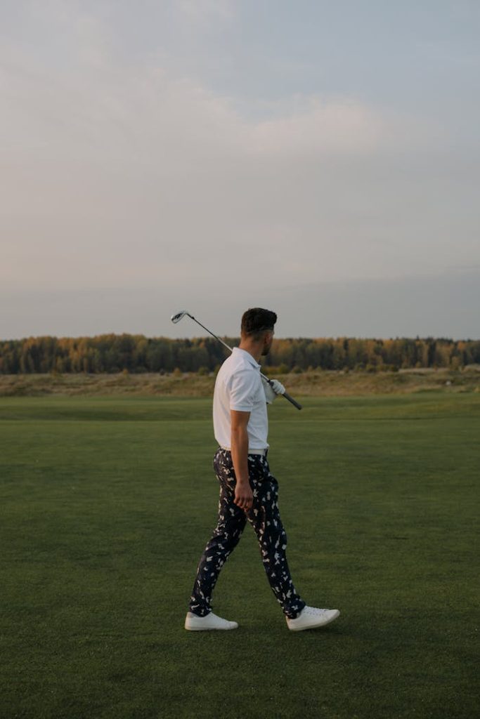 A stylish man walking on a serene golf course carrying a club over his shoulder.