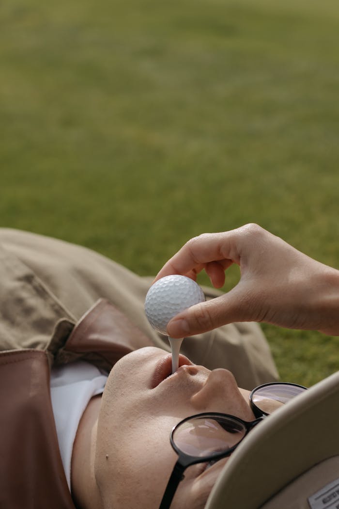 Artistic shot of a person lying on grass holding a golf ball on tee with mouth.